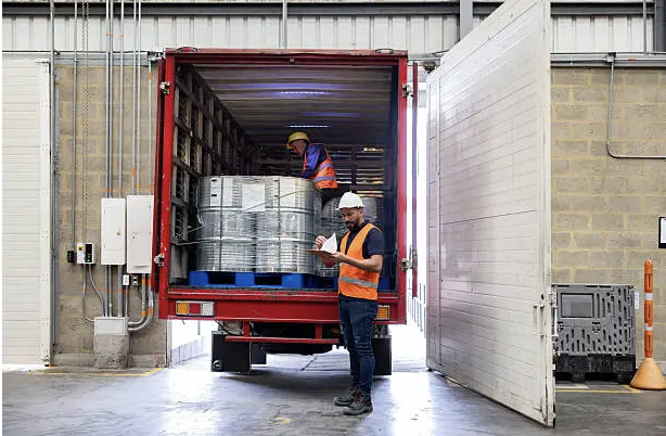 Logistics coordinator managing shipment documentation and coordinating with warehouse staff, loading dock area with shipping containers visible